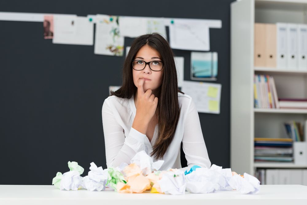 Young woman with writers block sitting in an office with a desk littered with crumpled paper as she sits looking thoughtfully into the air with her finger to her chin seeking new ideas